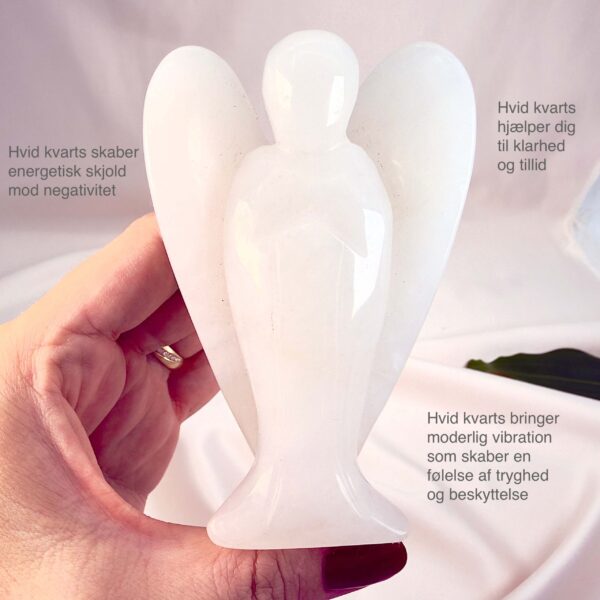 Hvid kvarts 10 cm krystal engel imellem fingre med hvid baggrund og grønt blad og tags