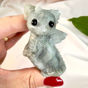 Labradorit krystal babydrage mellem fingre på hvid baggrubd med grønt blad