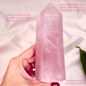 Rosakvarts krystal tårn mellem fingre med hvid baggrund og grønt blad og tags