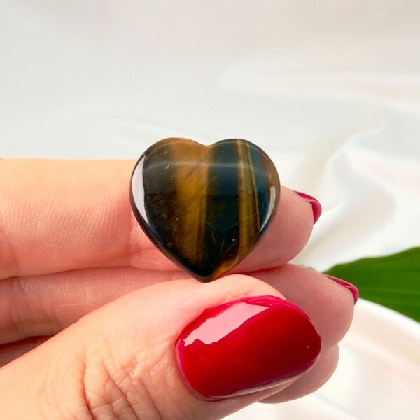 Tigerøje mini krystal hjerte mellem fingre med hvid baggrund og grønt blad